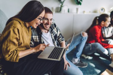 Neşeli genç kadın ve adamın yan görünüşü, diğer arkadaşlarıyla aynı odada oturup dizüstü bilgisayar kullanmaları ve komik videolar izlerken gülmeleri. 