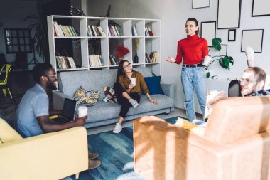 Bir grup güler yüzlü, çok kültürlü arkadaş rahat koltuklarda oturup gündelik giysiler giymiş, parlak odanın ortasında duran bayanları dinliyorlar.