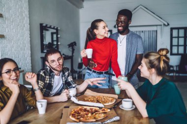 Çok ırklı genç hippiler lezzetli pizzalarla yuvarlak masa çiziyor sıcak içeceklerin tadını çıkarıyor ve şık bir evde birlikte eğleniyorlar.