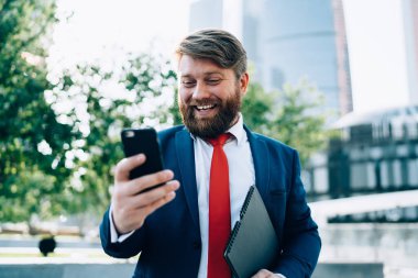 Zarif takım elbiseli, kırmızı kravatlı, akıllı telefon kullanarak partneriyle sohbet eden, modern şehrin parkında yürüyen neşeli, güler yüzlü adam.