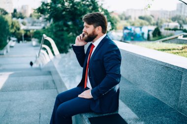 Şık takım elbiseli, şık ve kırmızı kravatlı, modern şehrin parkında çitin üzerinde oturan akıllı telefon kullanarak iş arkadaşlarıyla iş problemlerini tartışan stresli bir erkek.