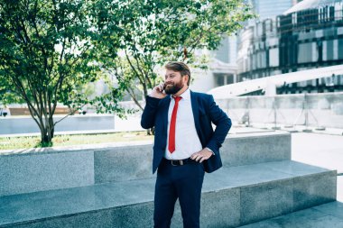 Koyu renk takım elbiseli, kırmızı kravatlı neşeli sakallı adam iş merkezinde cep telefonuyla konuşuyor. 