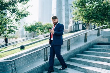 Sakallı, düşünceli, şık takım elbiseli, kırmızı kravatlı bir işadamı elinde cep telefonuyla merdivenlerden inip kameraya bakarken. 