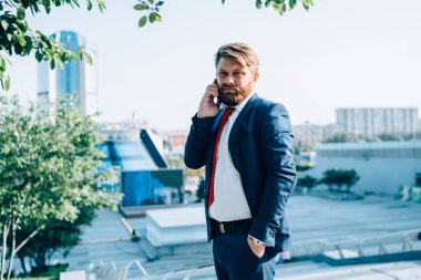 Şık takım elbiseli hoşnutsuz yetişkin erkek, güneşli bir günde iş yerinde iş arkadaşıyla cep telefonuyla konuşuyor.