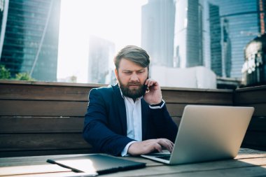 Mavi takım elbiseli konsantre sakallı iş adamı cep telefonuyla modern güneşli şehirde gökdelenli terasta konuşuyor.