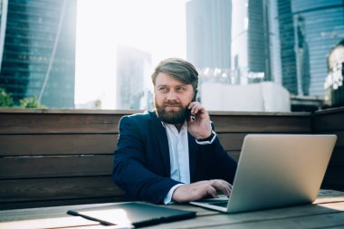 Mavi ceketli ve beyaz gömlekli sakallı adam şehir merkezindeki kafedeki ahşap masada oturmuş cep telefonu sörf laptopuyla konuşuyor.