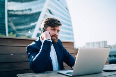 Ciddi odaklı, resmi takım elbiseli, cep telefonuyla konuşan, modern ayna gökdelenleriyle şehir merkezinde laptopuyla çalışan sakallı bir erkek.