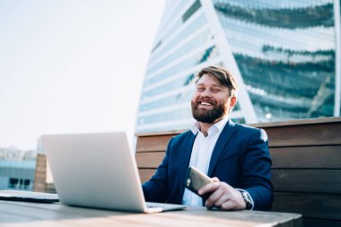 Şık takım elbiseli neşeli, modern bir genç elinde cep telefonu, elinde dizüstü bilgisayarla sokak kafesinde kameraya gülümsüyor.