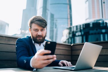 Modern resmi iş adamı elinde akıllı telefonu tutarken sokak kafesinde bilgisayarını masaya koyup kameraya bakıyor.