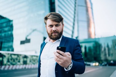 Resmi giyinmiş, kendine güvenen sakallı bir adam gün batımında bulanık gökdelenlere karşı sokakta akıllı telefonla iletişim kurarken kameraya bakıyor.