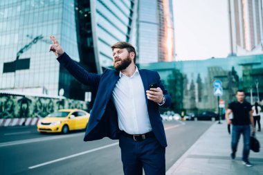 Sokakta dikilirken ve gece Metropolis 'teki bulanık gökdelenlere karşı akıllı telefon kullanırken kaldırılmış kollu taksiyle resmi giyinmiş kendine güvenen sakallı iş adamından.