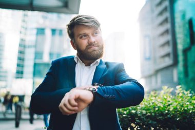 İş hayatı tarzında dalgın sakallı bir erkek ofis çalışanının kol saati kullanırken ve bulanık arka plana bakarken giydiği kontrol çizelgesinden. 