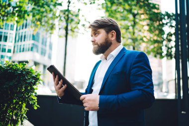 Kenarlarında ağaçlar ve ofis binalarıyla şehir merkezinde dikilirken elinde tablet olan düşünceli, kendine güvenen yönetici erkeğin yan görüntüsü.