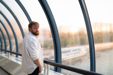 Karizmatik, ciddi, sakallı, beyaz gömlekli, kolları sıvalı bir erkek gün batımında yaya köprüsünde kameraya bakıyor.