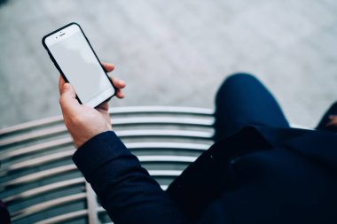 Yukarıdan gelen beyaz ekranlı, temiz cep telefonu elinde koyu renk elbiseli, asfalt zeminde oturan bir mahsul var.