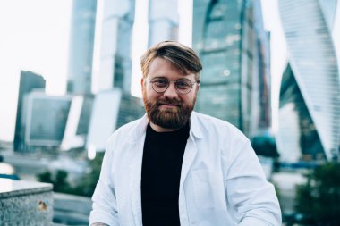 Kırmızı sakallı, beyaz tişörtlü ve siyah tişörtlü, arka planda ayna gökdelenleri olan mutlu, zeki bir erkek.