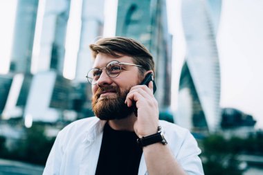 Akıllı, entelektüel, gözlüklü, gayri resmi giyinmiş, arka planda modern ayna şehri gökdelenleri olan cep telefonuyla konuşan kızıl sakallı erkek. 
