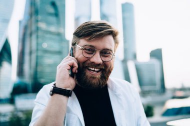 Şehir merkezinde cep telefonuyla dolaşırken kameraya gülümseyen optik gözlüklü neşeli erkek programcının portresini kapat. Cep telefonuyla iletişim kuran pozitif adam.