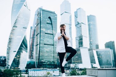 Arka planda şehir merkezindeki gökdelenlerle birlikte ağaç talimi yapan mutlu erkek portresi, dışarıda yoga eğitimi için Vrksasana yapan şık giyimli, pozitif bir adam.