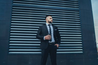 Kendine güveni tam sakallı bir adam, elinde içecekle, resmi giysilerin cebinde, modern binanın dışında dikilirken başka yere bakıyor.