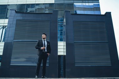 Çağdaş şehir caddesinde sıcak içeceğin tadını çıkarırken takım elbiseli, el ele tutuşmuş, gözlerini kaçıran dalgın sakallı bir erkek.