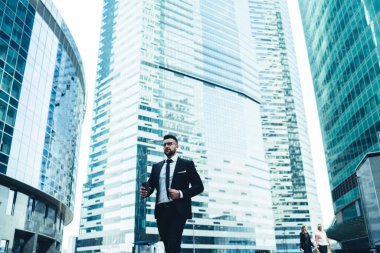 Şık siyah takım elbiseli, meşgul yetişkin işadamı şehir merkezinde cam cepheli, çağdaş, yüksek katlı ofis binalarına karşı cadde boyunca yürüyecek.