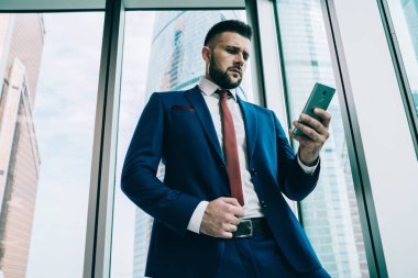 Şık takım elbiseli, düşünceli, sorunlu, ekranlara odaklanan ve akıllı telefonlu bir adamla gündüz vakti gökdelendeki ofis binasının penceresinin önünde duran bir adamdan.