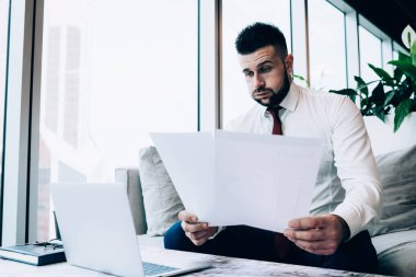 Resmi olarak modern erkek yönetici mali raporları okuyor ve ofis odasındaki pencerenin yanında oturup dizüstü bilgisayarla çalışırken iş sonuçlarından hayal kırıklığına uğruyor. 
