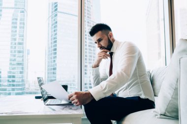 Ciddi bir erkek avukatın ofis odasındaki dizüstü bilgisayarla masada otururken dikkatlice kağıt okuması ve sözleşmenin detaylarını analiz etmesi. 