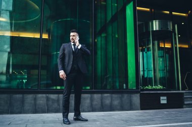 Siyah takım elbiseli ve kravatlı ciddi bir iş adamı telefon görüşmesi yapıyor ve önemli konuları tartışırken cam duvarlı modern binanın yanında duruyor.