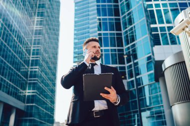 Güneşli bir günde gökdelenlerin ortasında dikilirken, elinde defterle bakan ve telefona cevap veren erkek patronun alçak açısı.