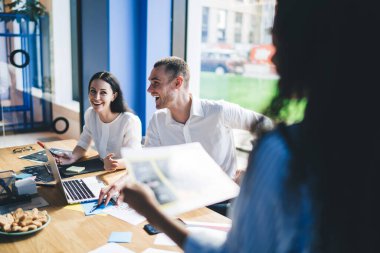 Çok ırklı erkek ve kadın çalışanlar, belgeler ve dizüstü bilgisayarlarla toplantı masasında oturuyor ve takımdaki yeni projeden bahsederken gülüyorlar.