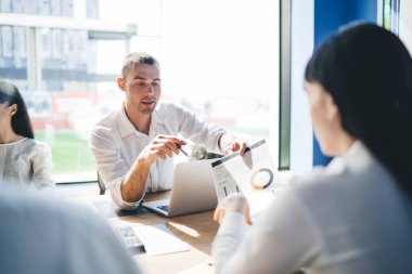 Ciddi iş adamlarının resmi giysiler içindeki meslektaşlarıyla raporları tartışırken, modern konferans odasındaki büyük ahşap masada gündüz pencerelerine karşı otururken yan görüntüsü