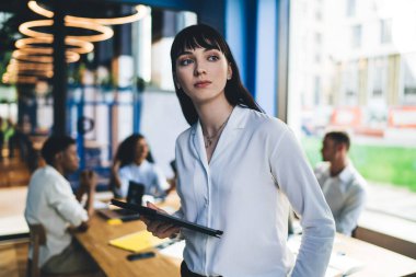 Beyaz, zarif tişörtlü, tabletli modern düşünceli esmer ve bulanık arka planda konuşan iş arkadaşlarıyla birlikte ofisten dışarı bakıyor. 