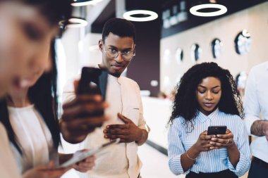 İnsanlar modern ofis lobisinde ayakta dikilirken ve akıllı telefonlar üzerinde sörf yaparken resmi gözlüklü neşeli Afrikalı Amerikalı adam fotoğraf çekiyor.