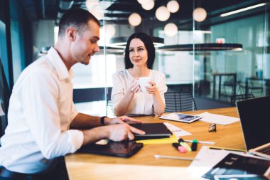 Ofis çalışanları modern konferans odasında ahşap masada aletlerle ve ofis malzemeleriyle tartışıyor ve bir fincan kahve içiyor.