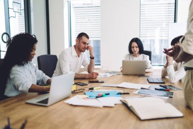 Yoğunlaştırılmış ofis çalışanlarından oluşan çok ırklı bir ekip toplantı sırasında dikkatle dinlerken dizüstü bilgisayarları ve belgeleriyle masanın önünde oturuyor.