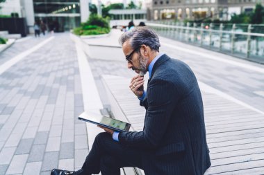 New York 'ta kenar mahallede bankta otururken modern bir aletle düşünülen temiz takım elbiseli, orta yaşlı düşünceli bir erkeğin yan görüntüsü. 