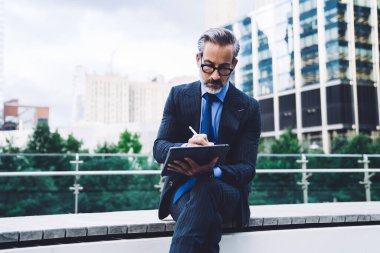 New York binalarının arka planında otururken Stylus 'la tablet kullanan konsantre orta yaşlı takım elbiseli ve gözlüklü bir adam.