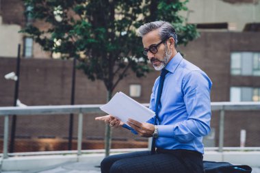 Takım elbiseli, gözlüklü, ahşap bankta oturan ve New York 'ta gazetelere bakan olgun bir adamın yan görüntüsü.