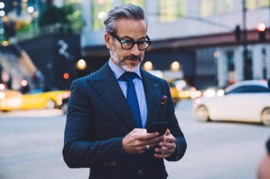 Yakışıklı, orta yaşlı, gözlüklü, takım elbiseli, telefonda yazan ve New York yoluna karşı duran bir erkek.