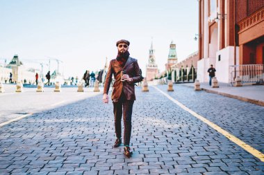 Güneş gözlüklü, kahverengi deri ceketli sakallı hippi şehir meydanında asfalt yolda yürüyor.