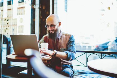 Genç içerikli, sakallı, gözlüklü, dizüstü bilgisayarlı, kafede gündüz vakti kahve içen kel adam.