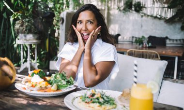 Şaşırtıcı yetişkin kadın yanaklarına dokunuyor ve çeşitli egzotik yiyeceklerle kafenin terasındaki ahşap masada otururken kameraya bakıyor.