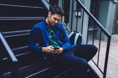 Kendine güvenen, kot gömlekli, sırt çantalı, merdivenlerde oturan ve New York caddesinde akıllı telefon kullanan yakışıklı adam.