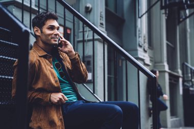 Moda ceketli genç yakışıklı adam New York caddesinde merdivenlerde oturuyor ve akıllı telefondan gülümsüyor.