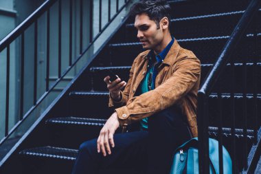 Modern dijital akıllı telefonlarda gezinen ve mesajlaşan gündelik giysiler içindeki ciddi meşgul erkek aşağıya bakıyor ve koyu demir merdivenlerde oturuyor.