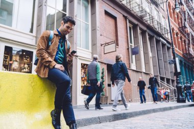 Günümüz akıllı telefonunda gezen ve mesajlaşan neşeli erkeğin altında otururken, gülümserken ve şehir arka planına bakarken