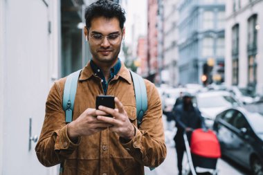 Gözlüklü, gülümseyen, kahverengi süet ceketli, sırt çantalı, cep telefonuyla mesajlaşan ve New York 'ta kaldırımda dururken müzik dinleyen yakışıklı İspanyol erkek.