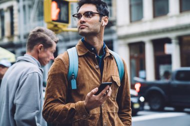 Gözlüklü, kahverengi süet ceketli, sırt çantalı ve kulaklıklı düşünceli bir İspanyol erkek New York caddesinde yeşil ışık beklerken müzik dinliyor ve başka tarafa bakıyor.